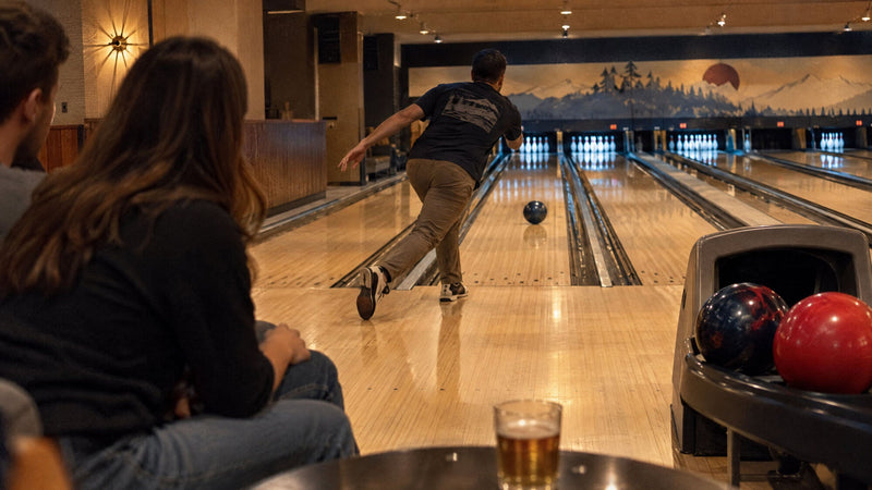 Gäste sehen einem Bowler beim Wurf zu, auf dem Kugelrücklauf liegen eine rote und eine blaue Kugel
