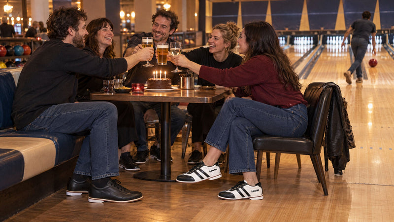 Venner skåler ved et fødselsdagsbord med kage i en bowlinghal, to af dem i BOWLIO Torino bowlingsko