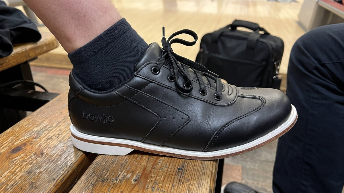 Foot in a black BOWLIO Nero bowling shoe on a wooden bench in a bowling center