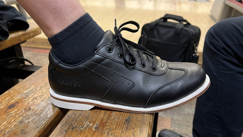 Foot in a black BOWLIO Nero bowling shoe on a wooden bench in a bowling center