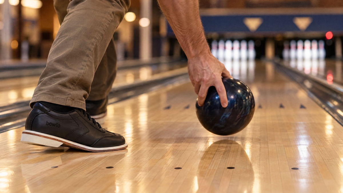 Hånd sætter en mørk bowlingkugle ned på banen ved foul-linjen, ved siden af en sort BOWLIO Nero-sko
