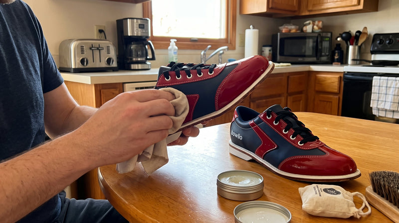 Person wiping a navy and red BOWLIO Verona bowling shoe with a soft cloth at the kitchen table, tins of leather balm beside it